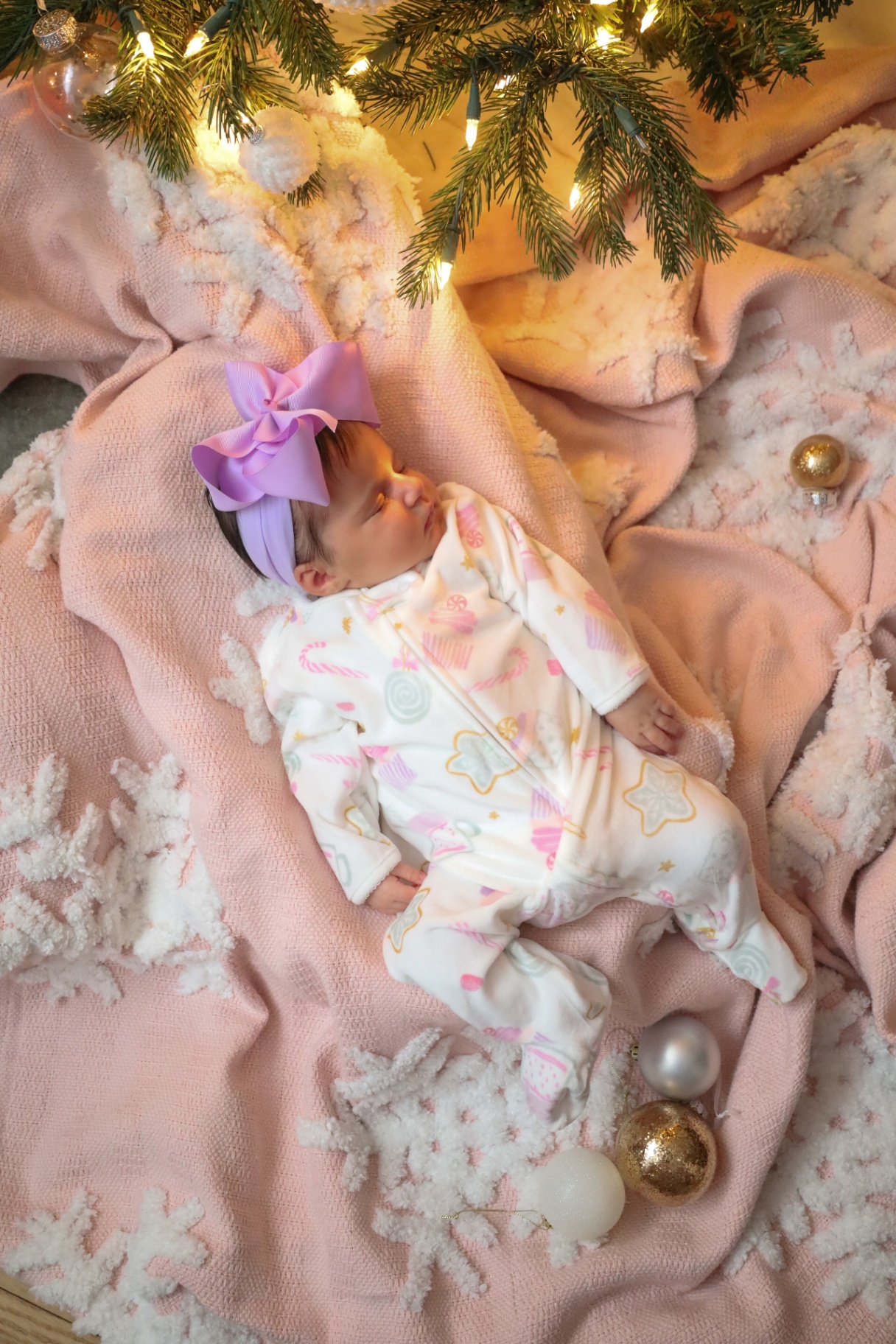 Newborn in pastel pajamas asleep on a blush blanket beneath the tree