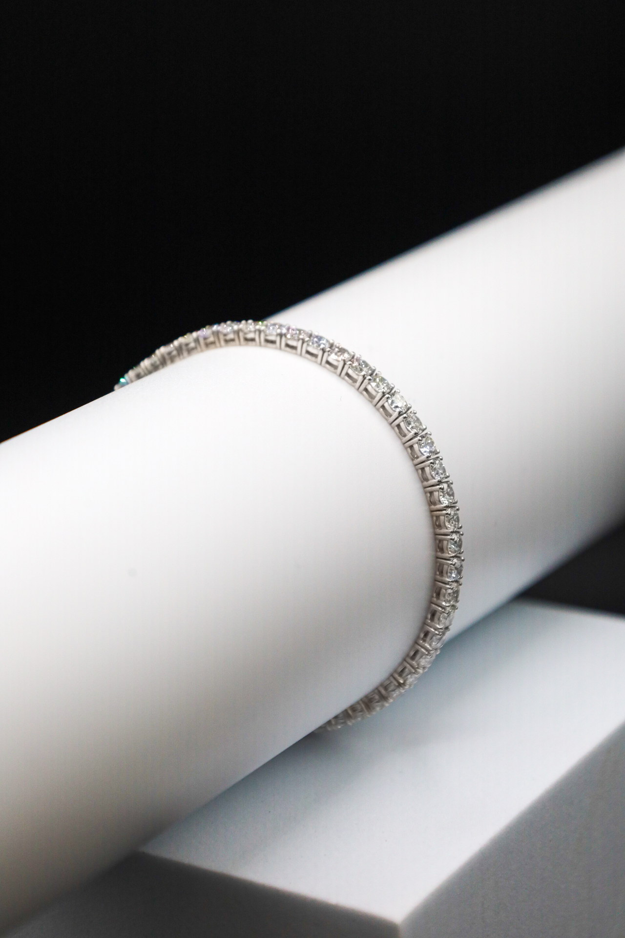 Round-cut diamond tennis bracelet displayed on a white cylindrical stand against a black backdrop