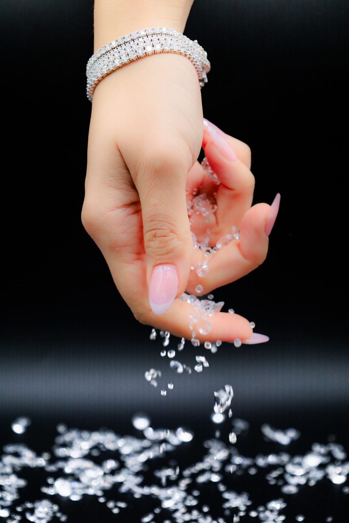 Hand pouring scattered crystals while wearing a stacked diamond bracelet against a black backdrop