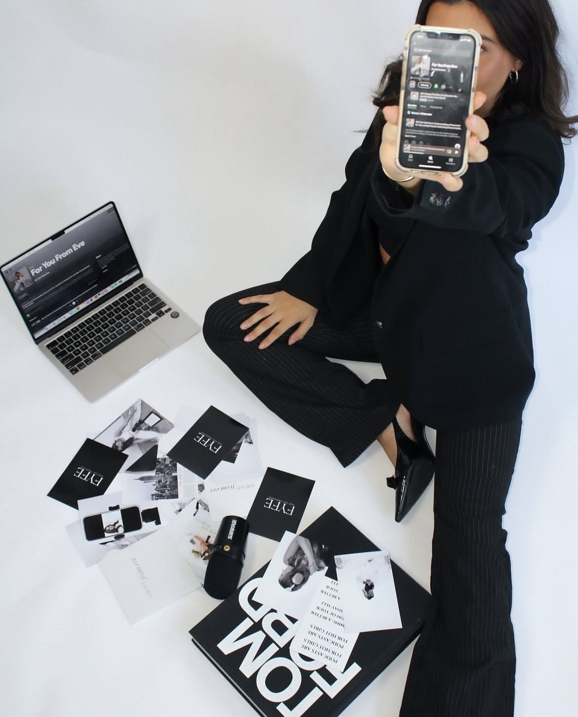 Brand founder seated on the floor holding up a phone beside branded print materials and laptop