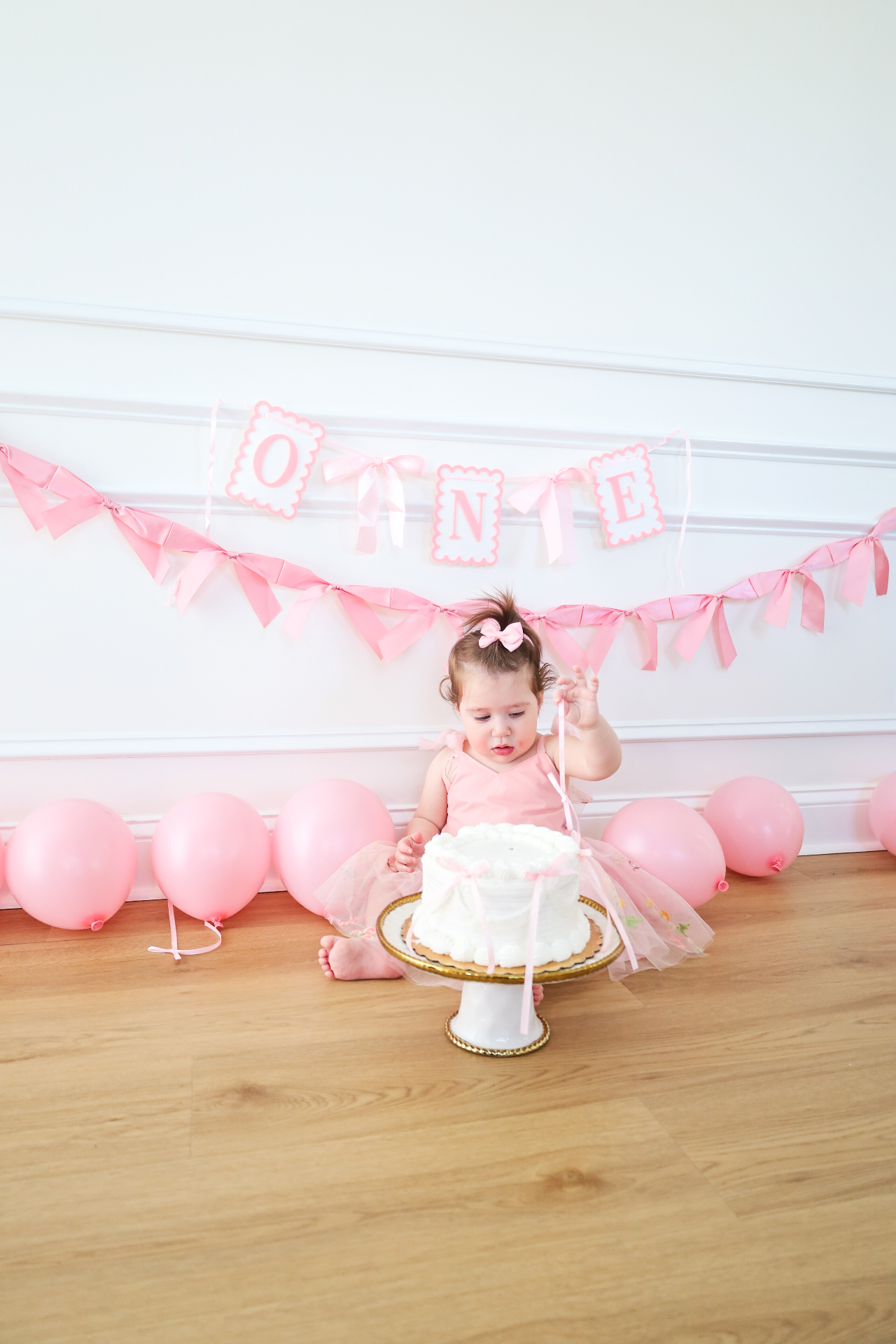 Baby girl reaching for a white birthday cake during a pink first birthday portrait session