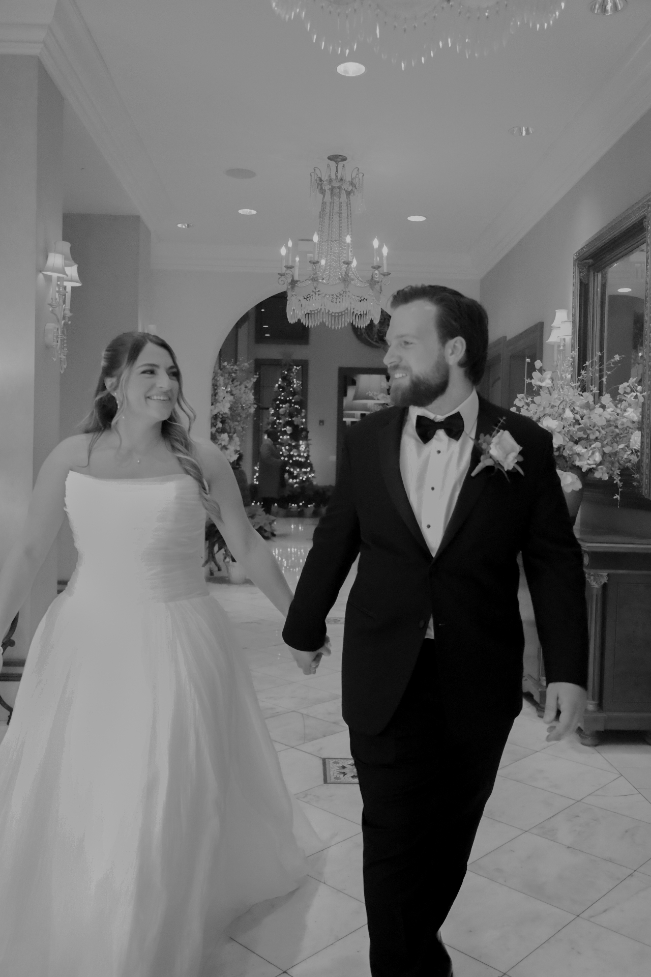 Black and white photo of the bride and groom holding hands walking through a chandelier-lit hall