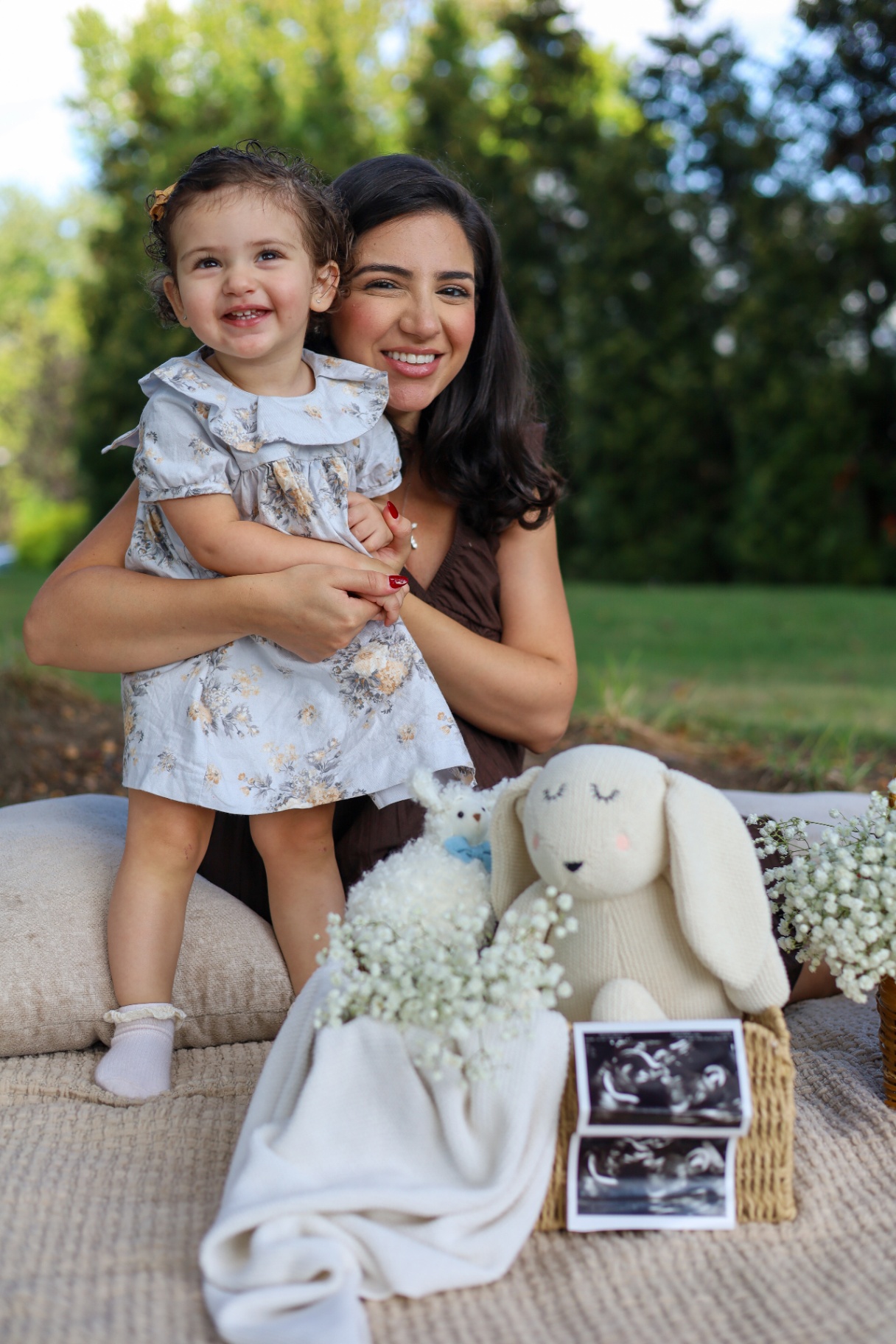 Mother holding her toddler daughter at an outdoor pregnancy announcement with ultrasound photos