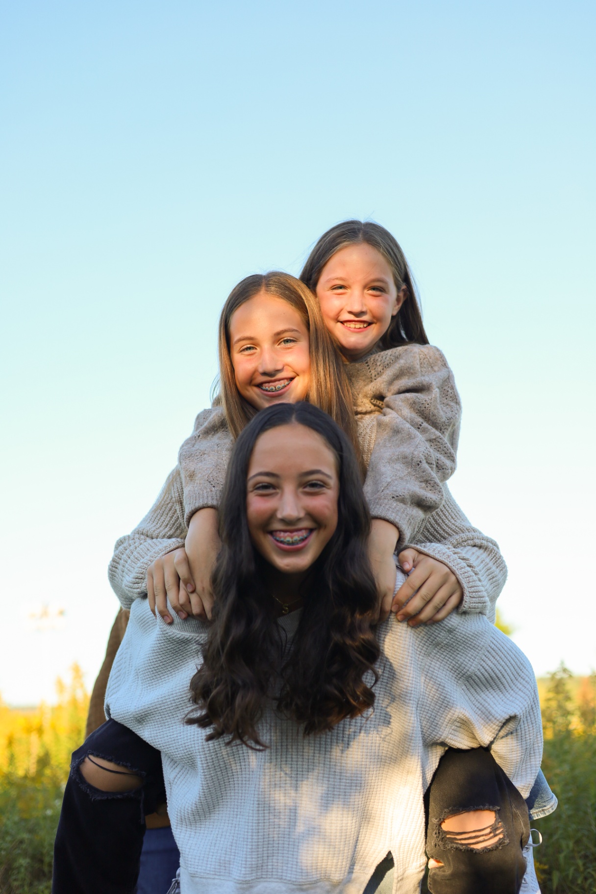 Three sisters piggyback-stacked and laughing in front of a golden sky