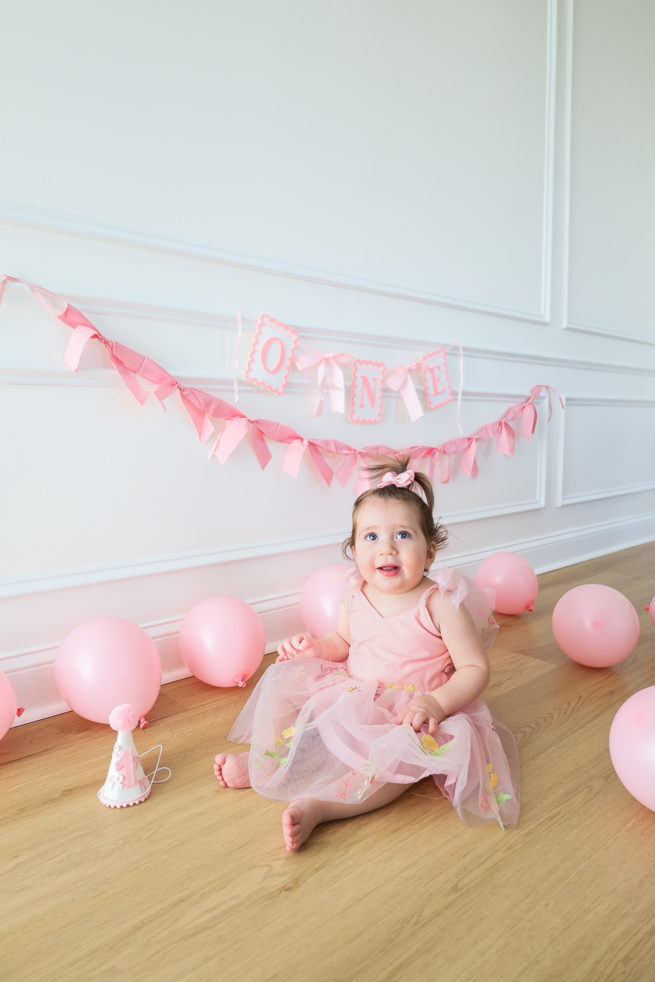 Baby girl in a pink dress sitting in front of a pink one birthday banner and balloons