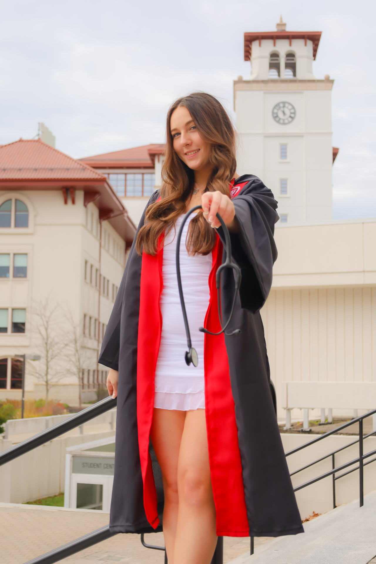 Nursing graduate in red-trimmed gown holding a stethoscope on campus steps