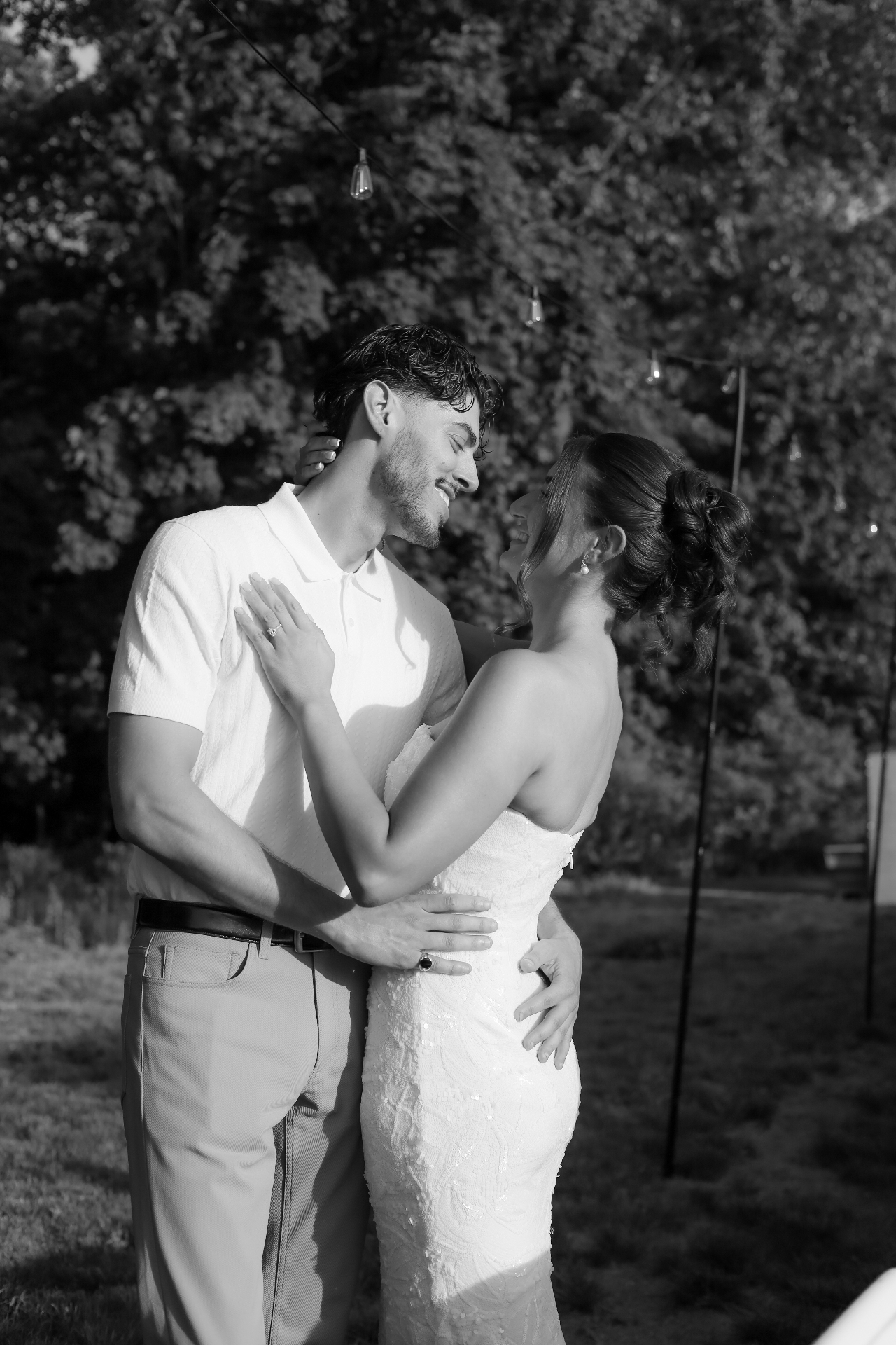 Black and white photo of the bride and groom sharing a tender embrace under string lights at golden hour
