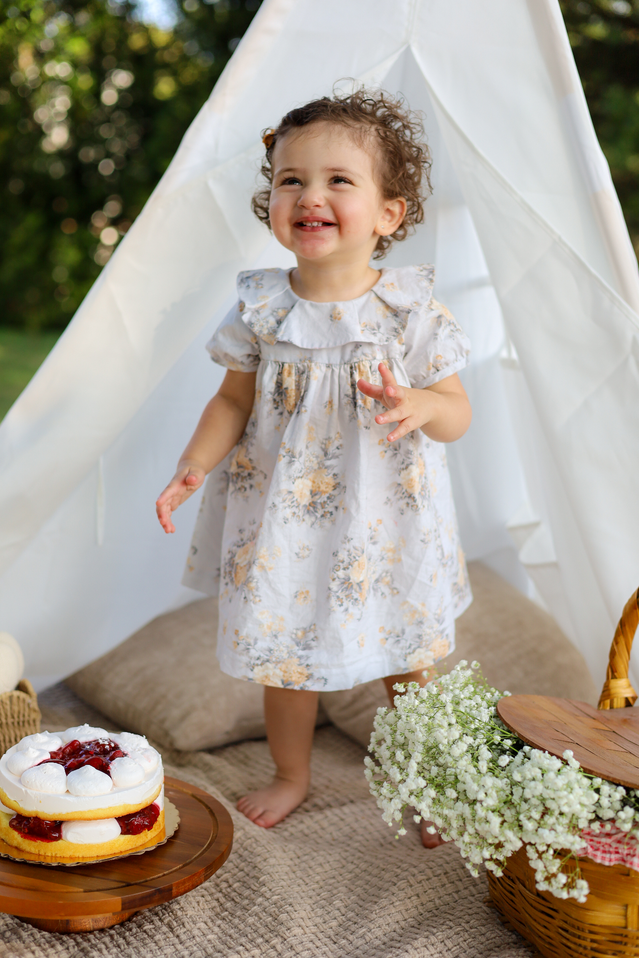Toddler in a floral dress smiling in front of a white teepee at an outdoor family session