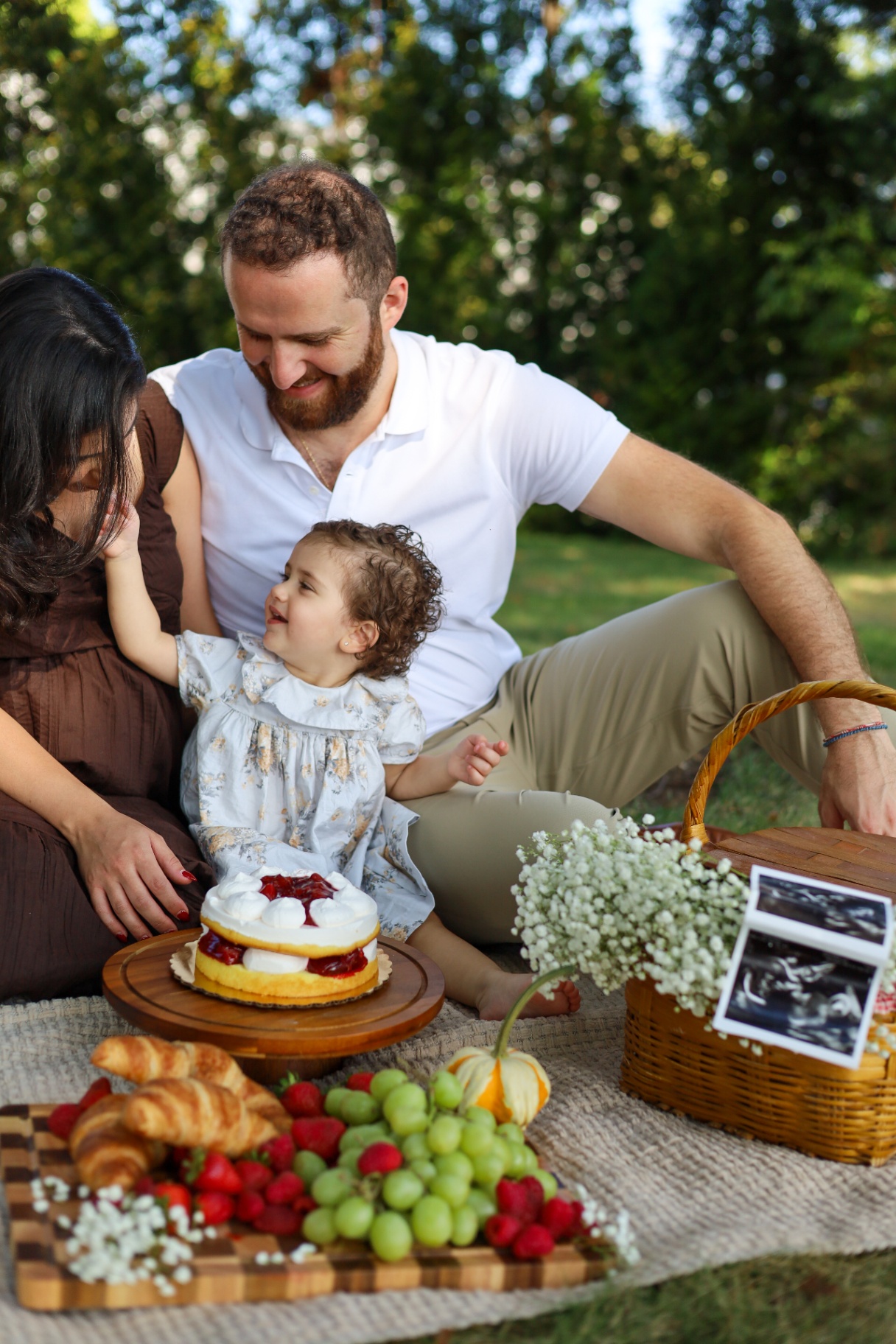 Family of three sharing a picnic with a strawberry cake and ultrasound photos
