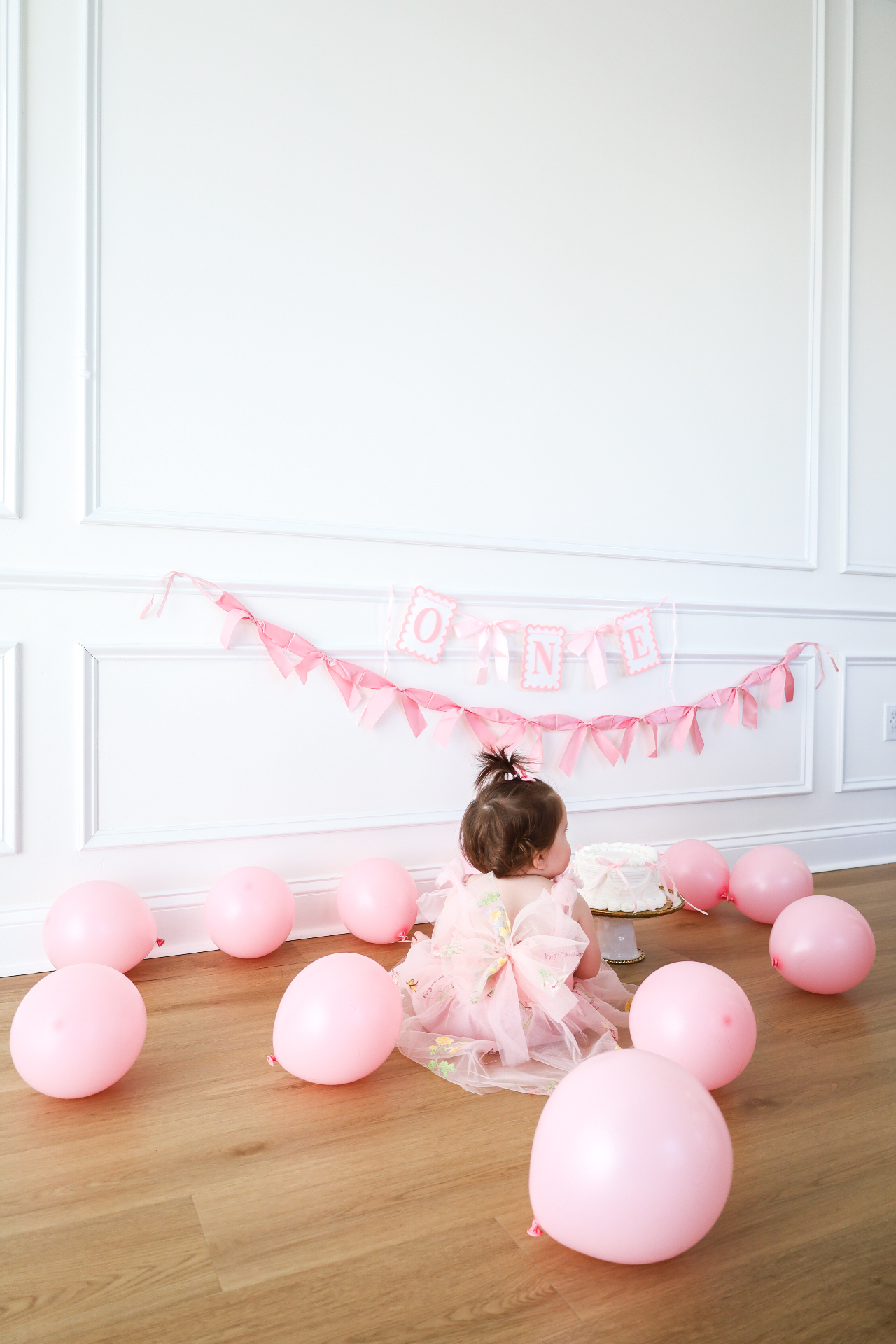 Back view of a baby girl in a birthday dress sitting among pink balloons facing her cake