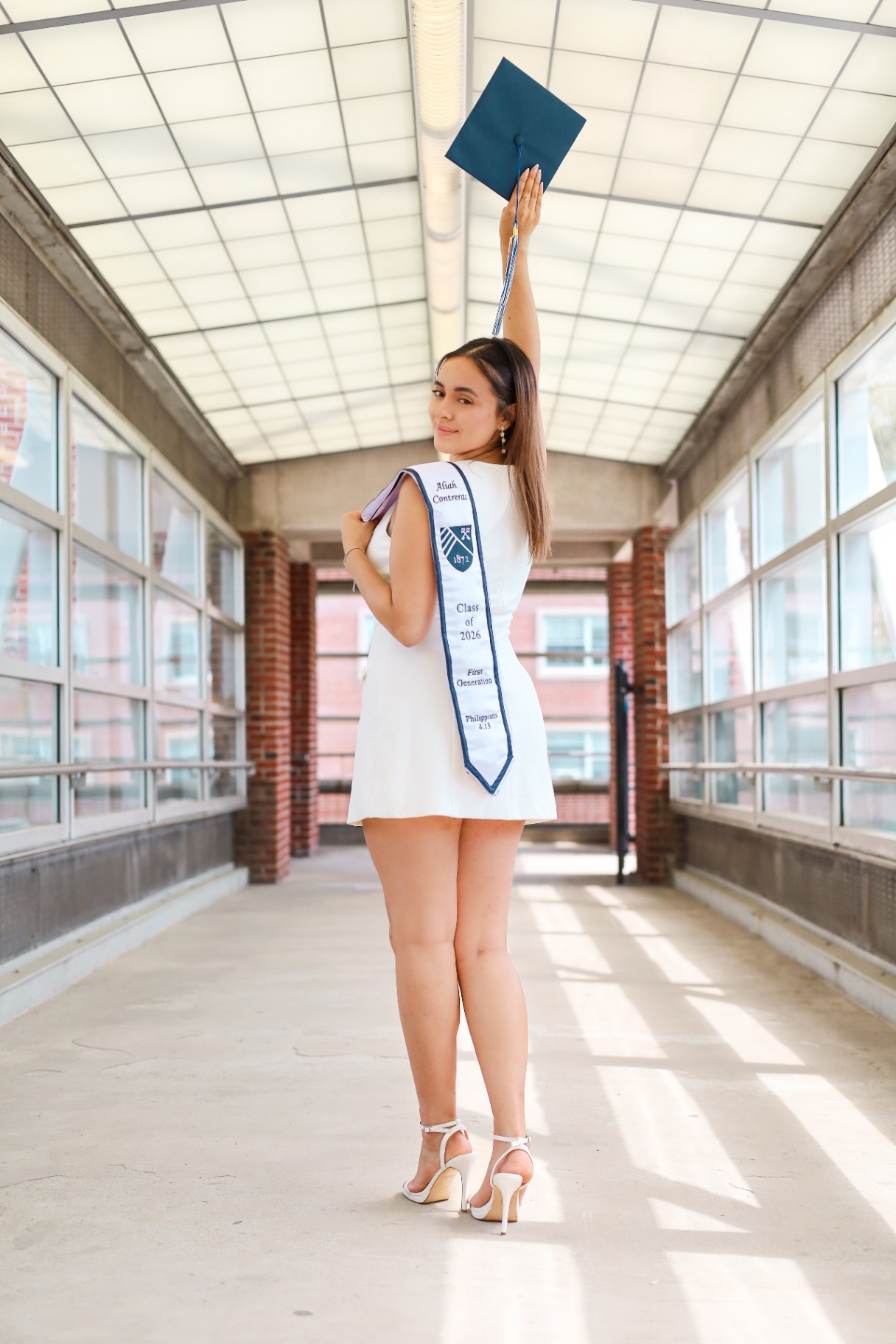 Graduate in white dress raising cap in a sunlit corridor