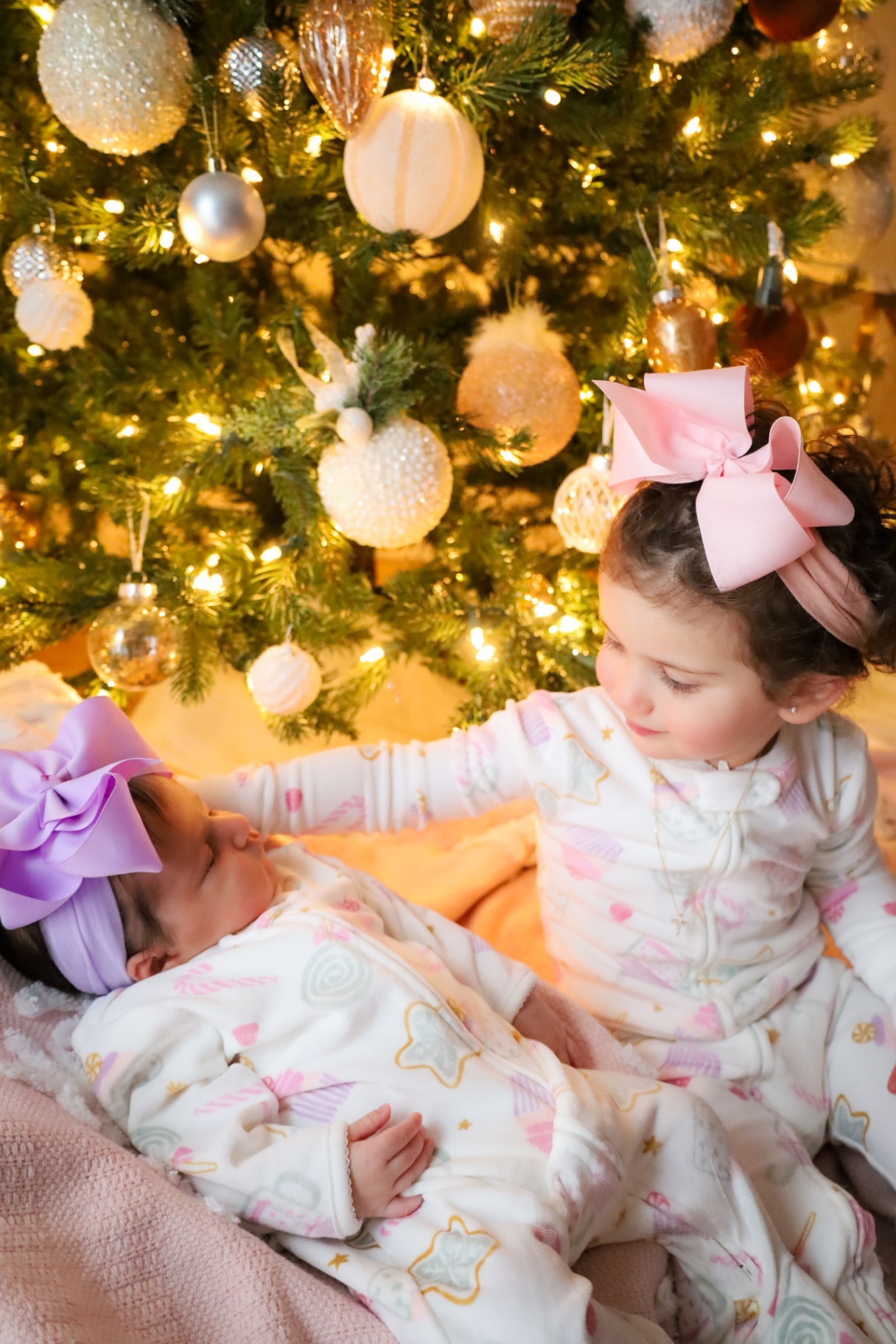 Big sister reaching toward her sleeping newborn sister by the tree