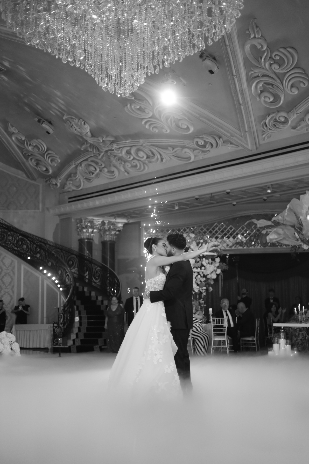 Black and white photo of the bride and groom sharing a kiss under a crystal chandelier with sparklers and low fog