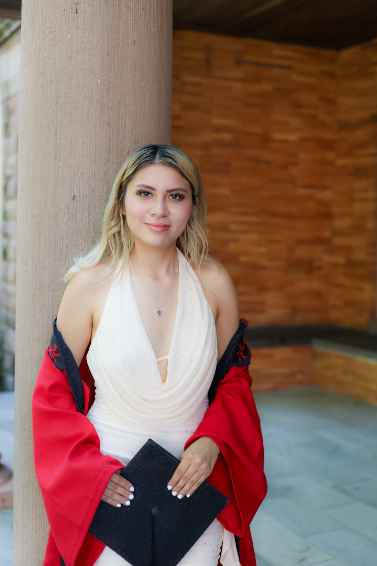Graduate in white dress leaning against a column with red gown draped