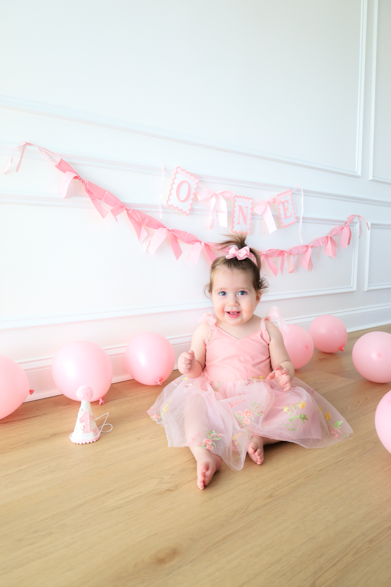 Smiling baby girl in a pink birthday dress surrounded by balloons and party decor
