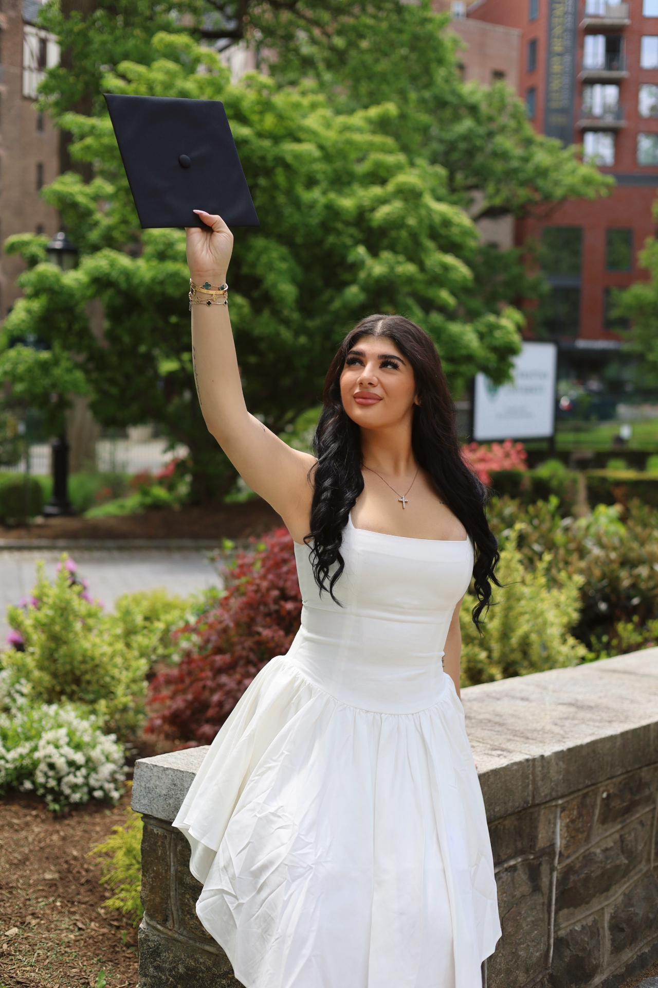 Graduate in a white dress raising her cap toward the sky in a city garden
